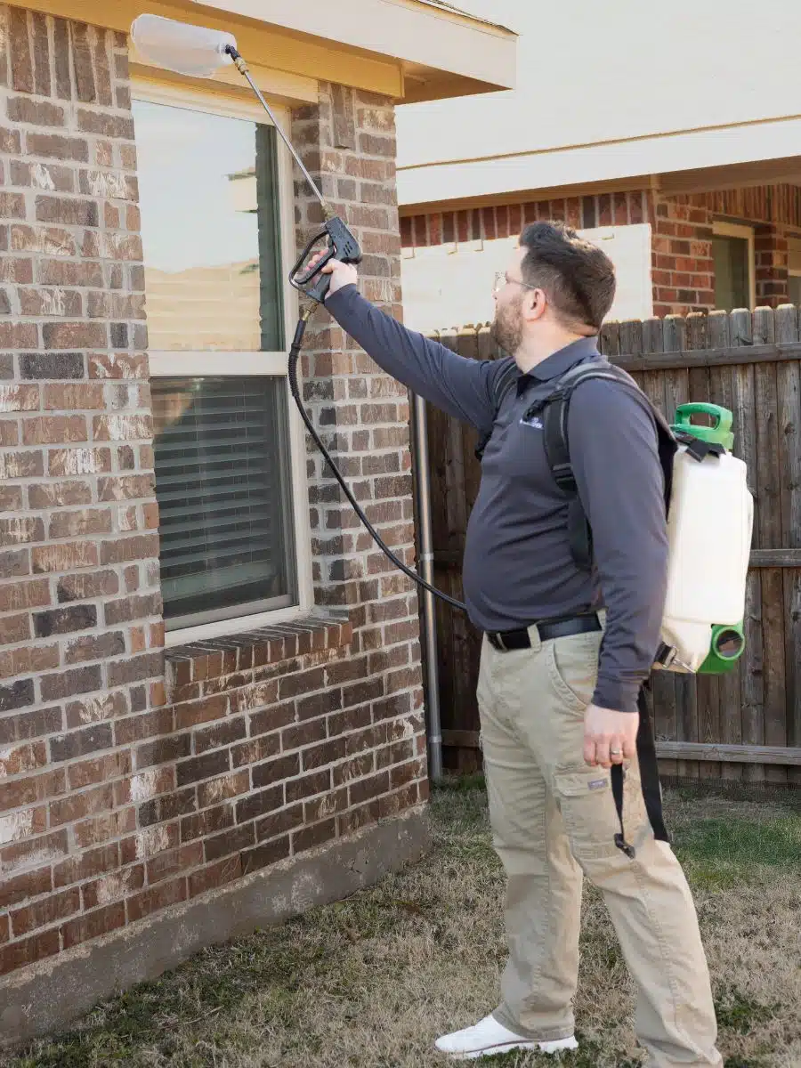 Preferred Pest Management technician performing wasp nest removal on a brick home in Richardson, TX using a backpack sprayer