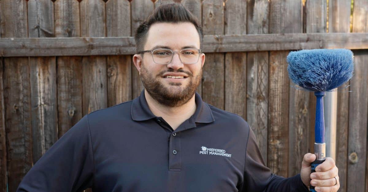 Preferred Pest Management technician holding a cobweb duster in front of a wooden fence in Sachse, TX