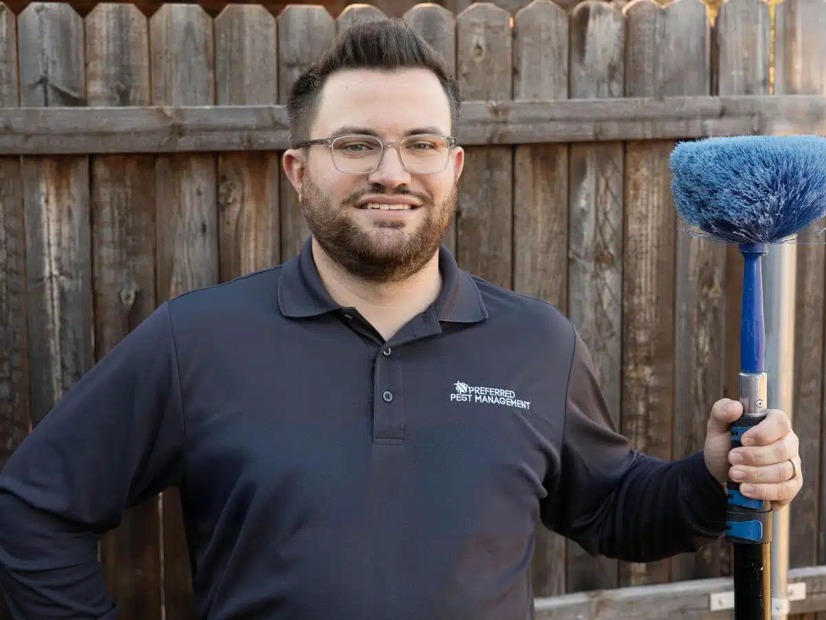 Preferred Pest Management technician holding a web removal tool in front of a wooden fence during a spider control service in Dallas