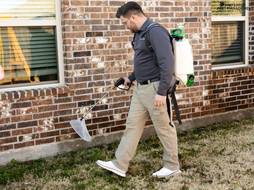 Technician applying perimeter ant treatment with a backpack sprayer along a Richardson TX home’s brick foundation